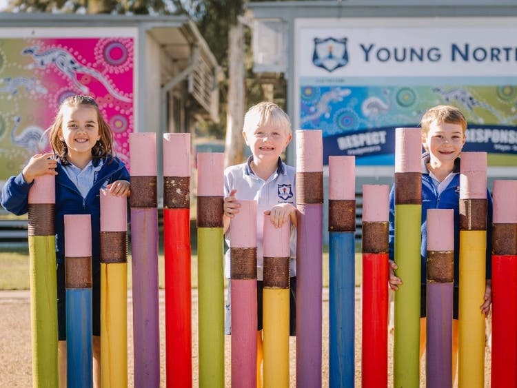 three students standing behide human sized coloured pencils